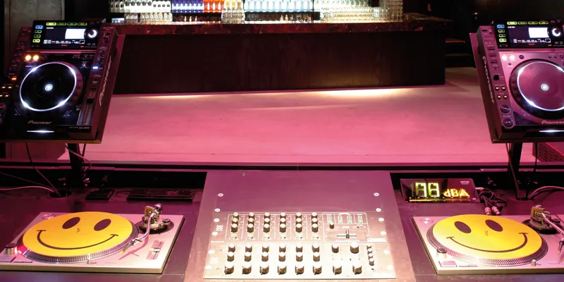 Club DJ booth with two Pioneer CDJ-2000 players on stands, two turntables and a mixer under pink light