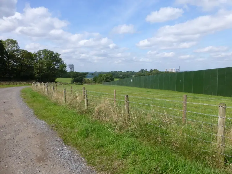 A gravel farm track beside a fenced field and the tall green screens around the Creamfields site near Daresbury