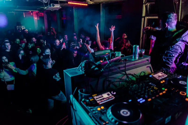 View from a DJ booth over a dark, crowded club dance floor, with the DJ setup in the foreground