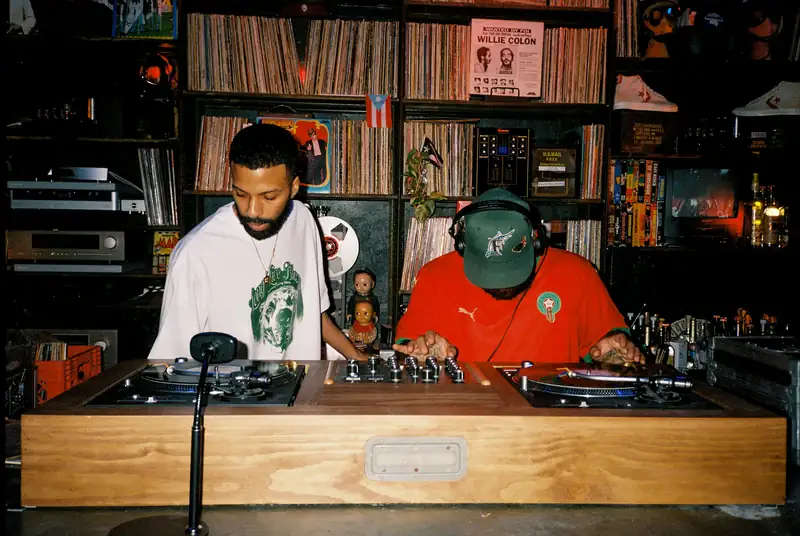 Two DJs mixing vinyl on turntables at a wooden console, with shelves of records behind them