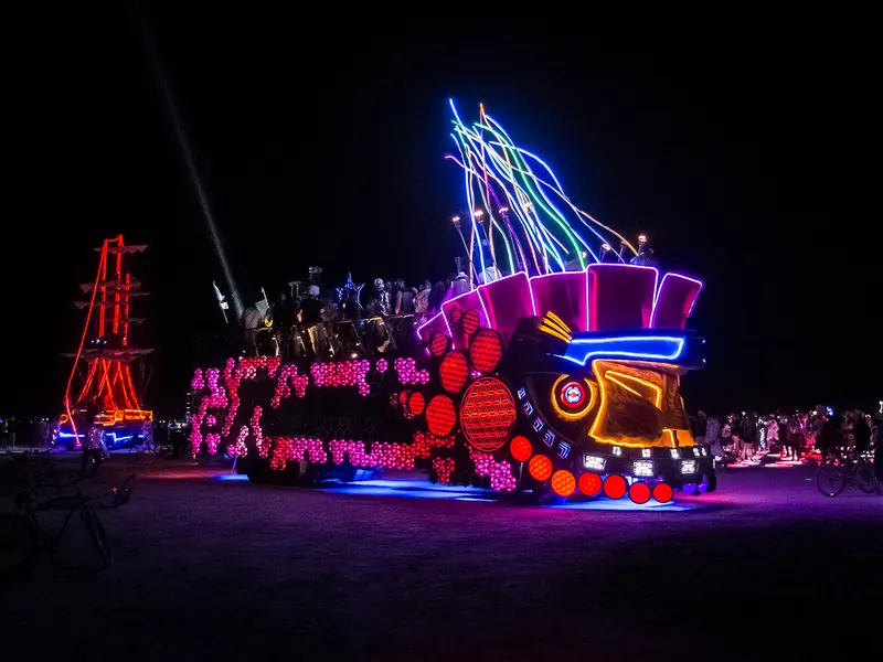 The Mayan Warrior art car lit up in neon at night in the desert, with people dancing on its top deck