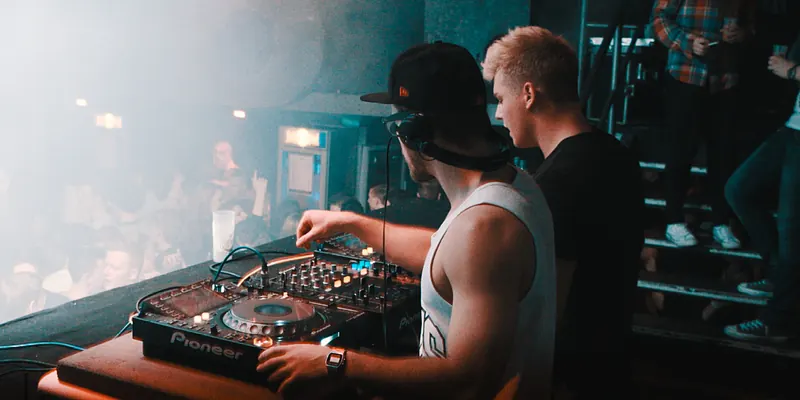 Two DJs in a club booth, one mixing on a Pioneer player and mixer while the other watches, above a dancing crowd