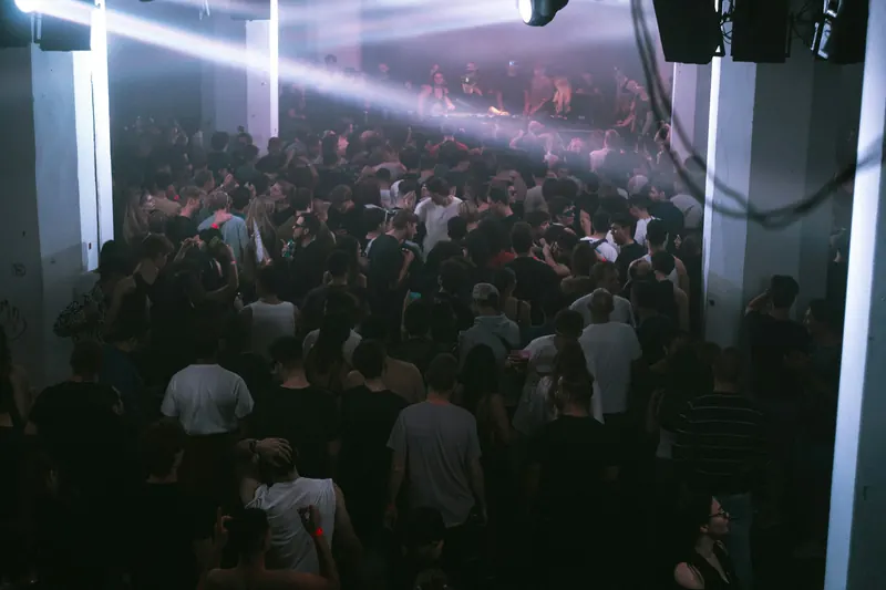 A packed club dance floor seen from the back of the room, with the lit DJ booth at the far end