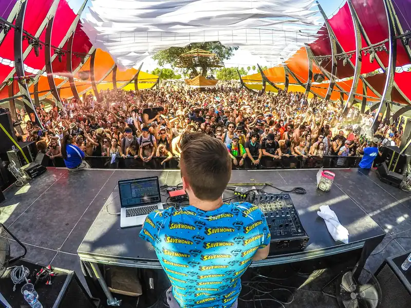 A DJ seen from behind on an outdoor festival stage, facing a big crowd at the barrier in the sun
