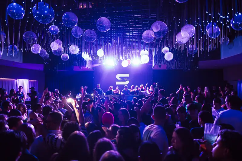 A packed club dance floor with raised hands in front of a DJ booth under purple lights and hanging glass spheres