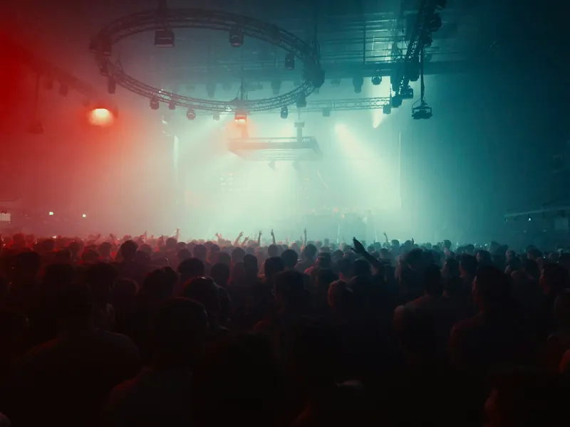 A packed crowd in a dark concert hall under red and blue stage lights and a ring of lighting trusses