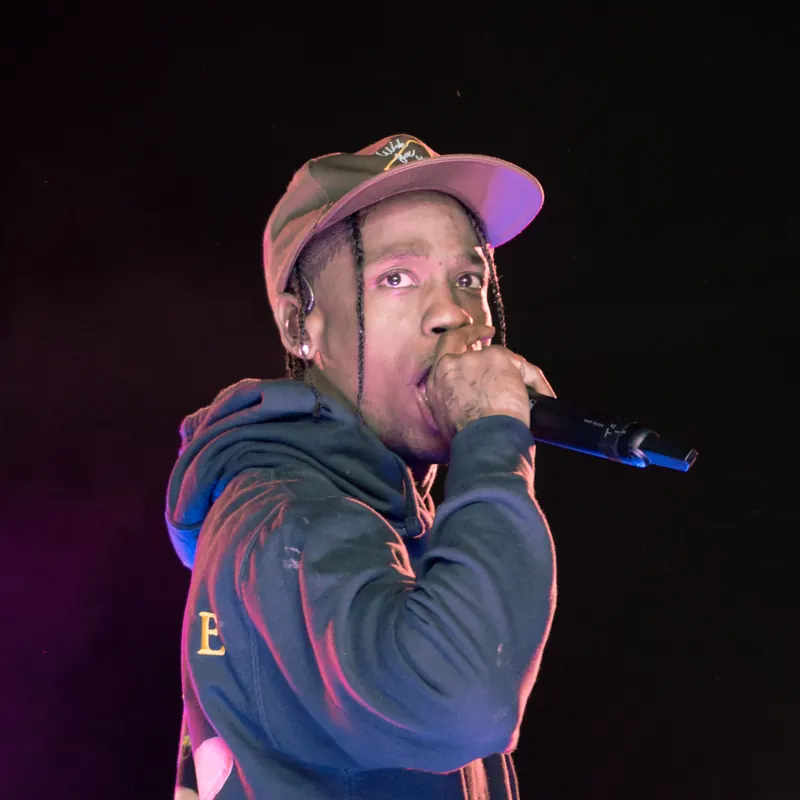 Travis Scott rapping into a microphone on stage, wearing a cap and a dark hoodie