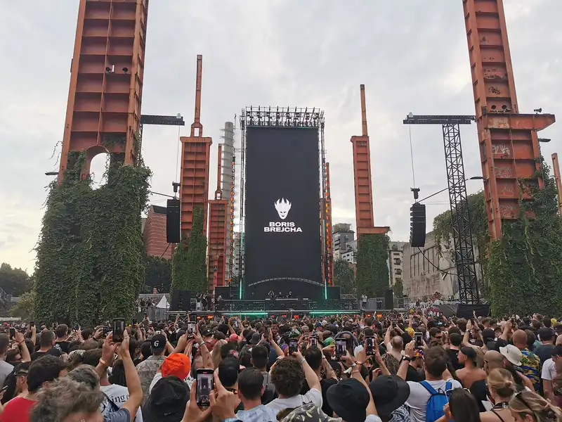 A festival crowd filming a tall stage screen that shows Boris Brejcha's name, between rusty industrial towers