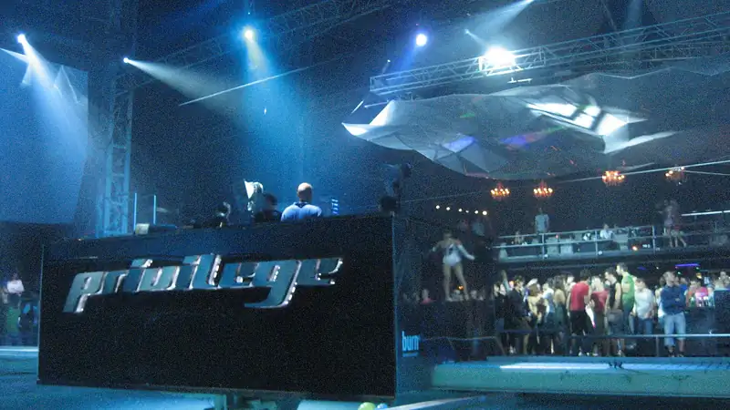 The DJ booth of Privilege Ibiza under blue stage lights, with dancers on the floor and a balcony behind it